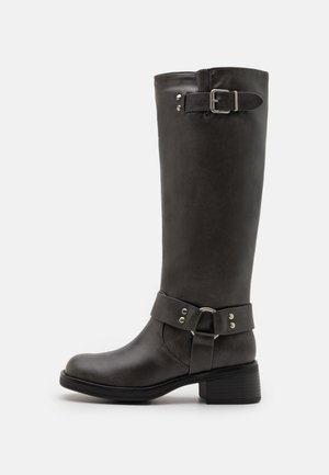 Botte haute en cuir marron foncé avec talon bas, bride décorative à la cheville avec anneau en métal et boucle réglable près du haut.