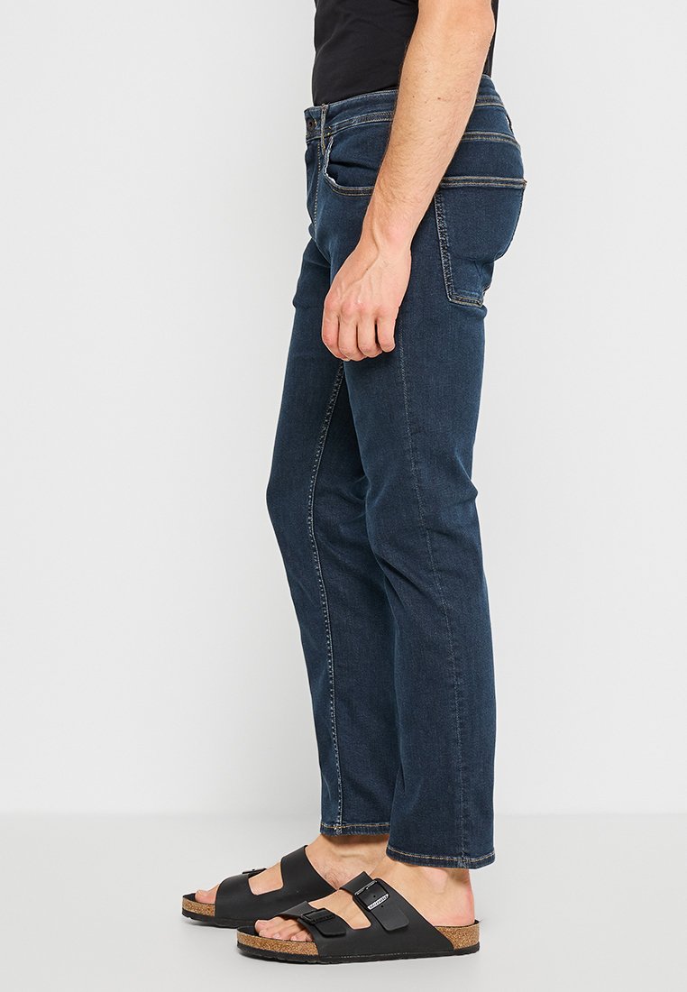 Dunkelblaue Jeans mit gerader Passform, die das traditionelle Fünf-Taschen-Design und kontrastierende Nähte aufweisen, getragen mit schwarzen Sandalen.