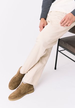 Person sitting on a chair wearing beige pants, brown suede shoes, and a long-sleeve checkered shirt, legs crossed at the ankles.
