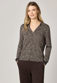 Femme portant un chemisier boutonné à manches longues à imprimé léopard et un pantalon foncé, debout les mains dans les poches sur un fond uni.