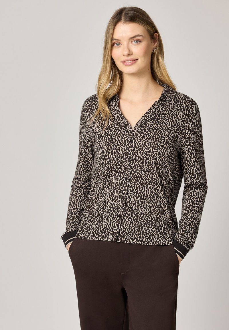 Femme portant un chemisier boutonné à manches longues à imprimé léopard et un pantalon foncé, debout les mains dans les poches sur un fond uni.