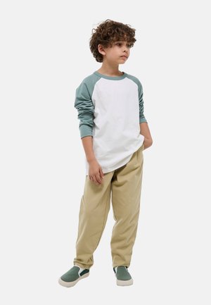 Garçon aux cheveux bouclés portant une chemise blanche et verte à manches longues, un pantalon beige et des chaussures vertes sans lacets, debout avec une main dans la poche.