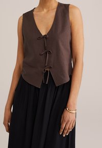 Brązowy top bez rękawów z trzema zapięciami na przodzie, noszony z czarną spódnicą i złotą bransoletką na prawej ręce.
