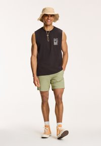 Sort ærmeløs skjorte med en grafisk lomme, grønne shorts, tan farvet bøttehat og orange sneakers mod en ensfarvet lys baggrund.