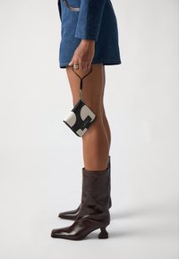 Svart og kremmønstret lommebok med glidelås og nøkkelring, holdt i hånden. En blå teksturert blazer og glansfulle brune ankelstøvler er synlige.