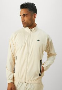 Man wearing cream-colored zip-up jacket with elastic cuffs and black-trimmed zip pockets, looking to the side against a plain background.