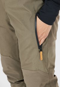 Pantalons vert olive avec une poche zippée et une tirette en cuir marron. La texture du tissu est lisse ; il présente des panneaux renforcés au niveau des genoux.