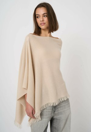 Kobieta w beżowym ponczo z frędzlami, nałożonym na jasnoszare dżinsy, stojąca na tle jednobarwnej białej ściany, patrząca w bok.