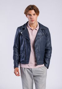 Chaqueta de motorista de cuero color marino con cierre de cremallera, cuello de solapa y dos bolsillos laterales, llevada sobre una camiseta polo rosa claro y pantalones grises.
