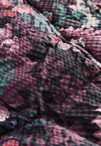 Texturierter Stoff mit dunklem Hintergrund und einem Blumenmuster in den Farben Lila, Pink und Grün, mit sichtbaren Nähdetails.