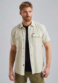 Beige Kurzarmhemd mit Knöpfen, zwei Brusttaschen, strukturiertem Stoff und einem schwarzen Unterhemd, das darunter sichtbar ist.