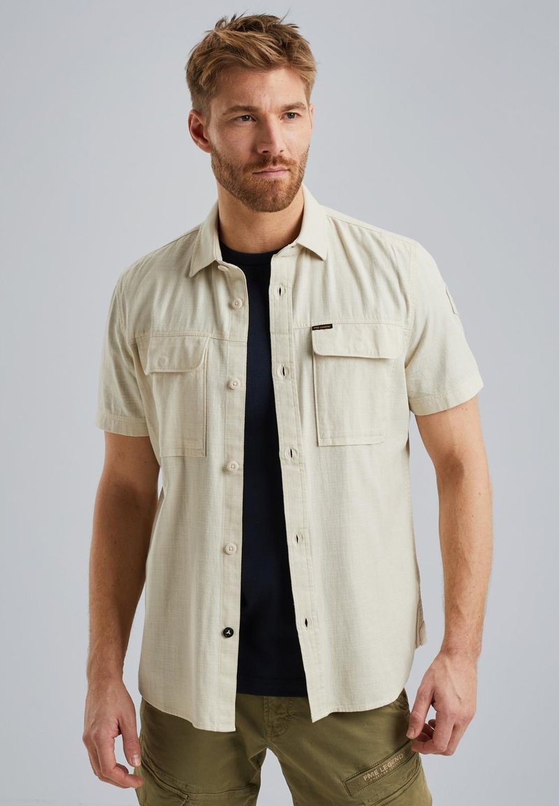 Beige Kurzarmhemd mit Knöpfen, zwei Brusttaschen, strukturiertem Stoff und einem schwarzen Unterhemd, das darunter sichtbar ist.