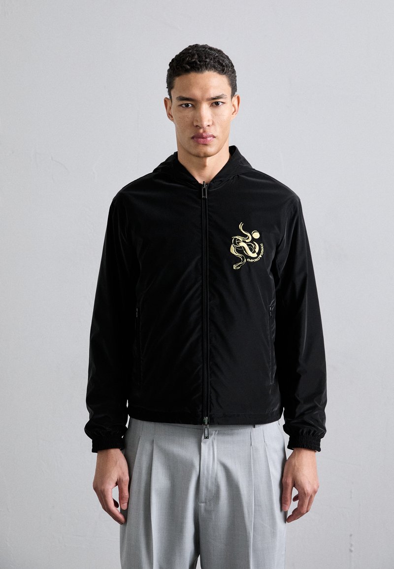 Casaco preto com fecho éclair e capuz, apresentando um design de polvo dourado no lado esquerdo do peito, punhos elásticos e bolsos laterais. Usado sobre calças cinzas claras.