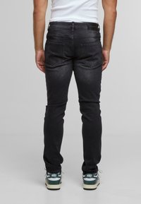 Persoon in donkere slim-fit jeans en witte Nike sneakers, staand met handen ontspannen naast het lichaam, gezien vanaf de achterkant.