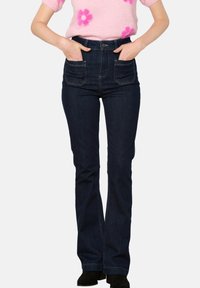 Mujer vistiendo jeans de tiro alto en azul oscuro con bolsillos frontales y botas negras, con las manos metidas en los bolsillos, sin rostro visible.