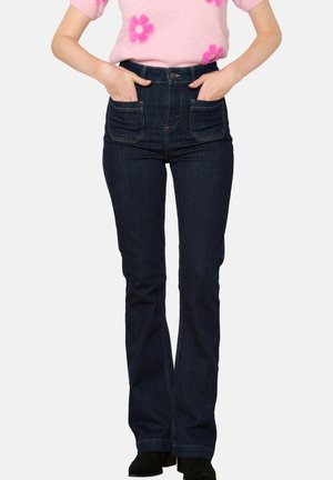 Mujer vistiendo jeans de tiro alto en azul oscuro con bolsillos frontales y botas negras, con las manos metidas en los bolsillos, sin rostro visible.