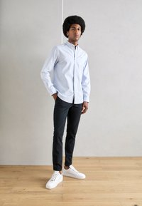 Lysblå stribet skjorte med knapper, sorte bukser, hvide sneakers; model står på trægulv med hænderne i lommerne, neutral baggrund.