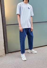 Jasnoszary, oversize'owy t-shirt z bawełny z małą niebieską grafiką, w zestawie z niebieskimi dżinsami i białymi tenisówkami za kostkę.