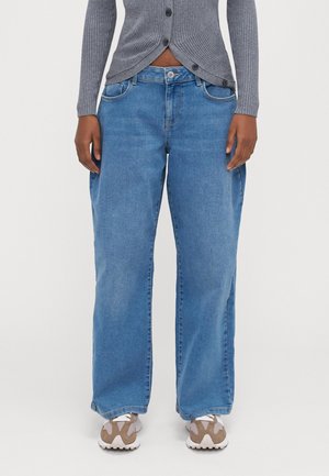 Person iført lyseblå vide jeans, grå ribstrikket cardigan med knapper og hvide sneakers med beige detaljer, stående mod en hvid baggrund.