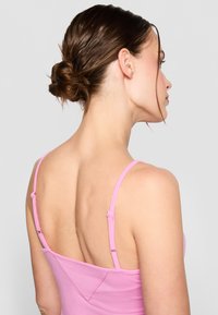 Kvinne med brunt hår i en lav hestehale iført en rosa singlet, vist fra bak og vendt litt mot høyre mot en grå bakgrunn.