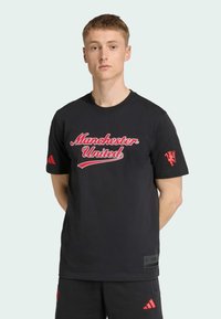 T-shirt en coton noir affichant "Manchester United" en écriture rouge, avec des bandes Adidas rouges sur les manches et un patch logo sur l'ourlet.