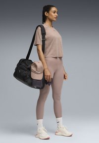 Μαύρη τσάντα duffel με ρυθμιζόμενο ιμάντα, ροζ καπέλο, γκρι πετσέτα και εφαρμοστά, ανοιχτό ροζ κολάν. Μοντέλο με γκρι τοπ και αθλητικά παπούτσια.