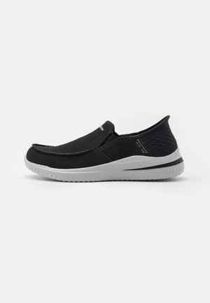 Zapato negro sin cordones con un upper texturizado, que presenta una puntera redonda, suela blanca y una mínima marca en el lado. Diseño ligero.