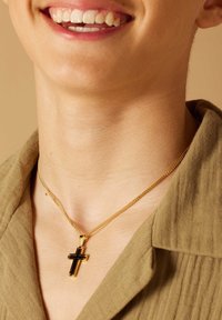 Gouden ketting met een zwarte kruispendant, met een glanzend oppervlak en textuur links, gedragen over een khaki overhemd met kraag.
