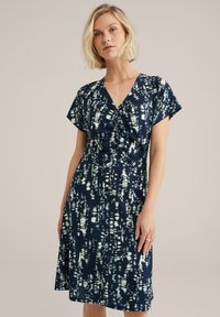 Robe marine longueur genoux avec des manches courtes, présentant un tissu texturé et un motif abstrait blanc et vert. Décolleté en V et détail froncé à l'avant.