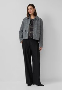 Grau strukturierte Jacke mit Kragen, zwei vorderen Taschen, mit Knöpfen geschlossen, kombiniert mit einem schwarzen Oberteil und lockeren schwarzen Hosen.