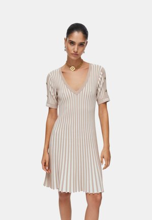 Kobieta ubrana w sukienkę w pionowe paski w kolorze beżowym i białym, z krótkimi rękawami i głębokim dekoltem w serek, ozdobiona złotym naszyjnikiem w kształcie kwiatów.