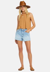 Brown ribbed sleeveless top paired with light blue denim shorts, featuring frayed hems. The model wears a brown woven straw hat and brown slip-on shoes.