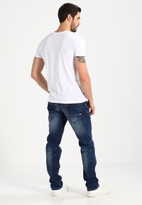 Witte t-shirt met korte mouwen in combinatie met donkerblauwe denim jeans. De jeans hebben achterzakken en een lichte vervaging. Model draagt witte sneakers.