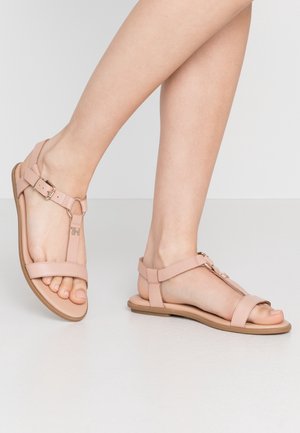 Jambes nues portant des sandales plates rose clair avec des brides à la cheville et de petits détails de boucle en métal, debout sur un sol blanc lisse.