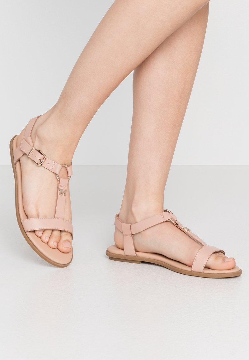 Jambes nues portant des sandales plates rose clair avec des brides à la cheville et de petits détails de boucle en métal, debout sur un sol blanc lisse.
