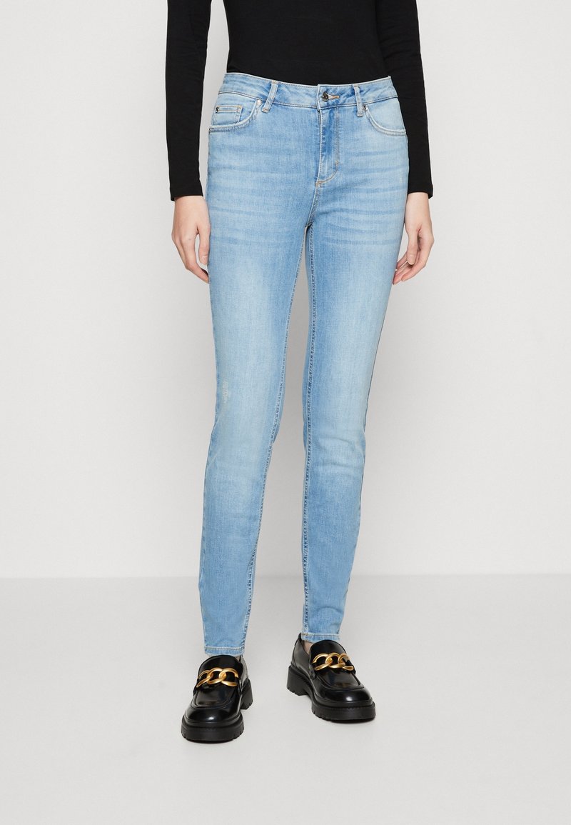 Personne portant un jean skinny bleu clair, un haut noir à manches longues et des mocassins noirs avec un détail chaîne dorée, debout sur un fond blanc.