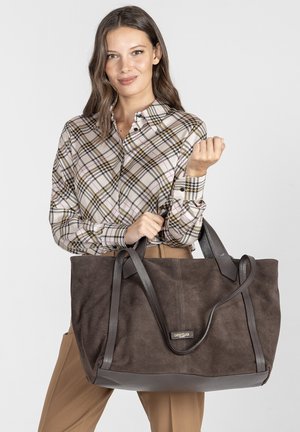 Brązowa zamszowa torba tote z ciemnymi skórzanymi akcentami i rączkami, charakteryzująca się pojemnym designem. Torba jest trzymana przez osobę ubraną w koszulę w kratę.