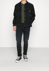 Camisa de botones a rayas negras y marinas, combinada con una camiseta verde, jeans negros y zapatillas altas. Diseño casual con bolsillos frontales.