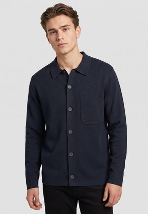 Junger Mann trägt eine dunkelblaue Strickjacke mit Knöpfen und einer Brusttasche sowie eine schwarze Hose, steht vor einem schlichten hellen Hintergrund.