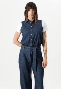 Denim jumpsuit met korte mouwen, donkere blauwe stof, knoopsluiting aan de voorkant en een gestrikt middel. Beschikt over een kraag en een losse pasvorm.