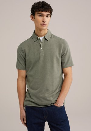 Camisa polo de manga corta en khaki con tejido texturizado, tapeta de tres botones y un cuello clásico. Combinada con pantalones de mezclilla oscuros.