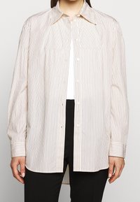 Valkoinen pitkähihaista napillista paitaa ohuilla vaaleanruskeilla pystysuorilla raidoilla. Paita sisältää klassisen kauluksen ja kaksi rintataskua; rento malli.