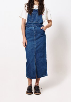 Femme portant un t-shirt blanc et une longue robe salopette en denim bleu avec une fente à l'avant, chaussures noires à lacets, debout devant un fond blanc.