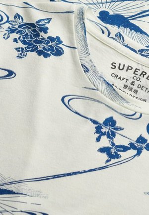 Crèmekleurig T-shirt met blauwe bloempatronen en golvende lijnen. De ronde halslijn en de stof hebben een zachte, gladde textuur.