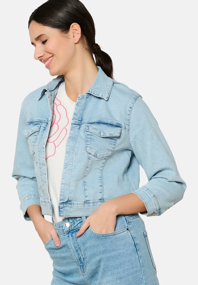 Hele sinine teksajakk lühikese disainiga, kahe esi-taskuga, nööpidega sulgemisega ja rullitud varrukatega, seljas valge särk roosa mustriga.