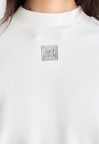 Primer plano de un suéter blanco con cuello acanalado, con un pin plateado cuadrado del logo "HUGO" centrado en el pecho.