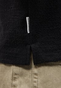 Κοντινή λήψη μαύρου υφασμάτινου ρούχου με υφή, με πλαϊνή σχισμή και λευκή ετικέτα που γράφει "Marc O'Polo DENIM" πάνω από μπεζ παντελόνια.