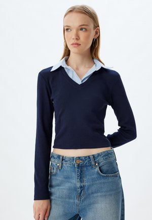 Jonge vrouw met lang blond haar die oorbellen draagt, een marineblauwe trui met V-hals over een blauw gestreept overhemd met kraag en een blauwe spijkerbroek.