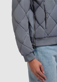 Grijze gewatteerde bomberjack met ribgebreide manchetten en tailleband, voorzien van een zilveren rits en een merklabel op de mouw. Denim jeans zichtbaar.