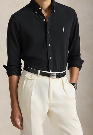 Uomo che indossa una camicia nera a maniche lunghe con bottoni e logo bianco, infilata in pantaloni color crema a vita alta con cintura nera e orologio al polso.